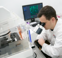 The photo shows a scientist in an apron observing a slide under a microscope. The monitor behind the scientist shows a confocal microscopy image showing the cells.