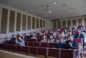 Zdjęcie przedstawia osoby zgromadzone na sali podczas jednego z wystąpień. Uczestnicy siedzą w rzędach na audytorium i klaszczą, dziękując prezentującemu za wystąpienie